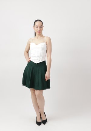 Kvinde med sort hestehale iført en hvid ærmeløs top, mørkegrøn plisseret nederdel og sorte hæle, der står mod en ensartet lys baggrund.