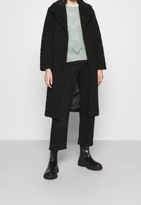 Manteau noir texturé avec un col châle, porté sur un pull en tricot vert menthe, associé à un pantalon noir et des bottines noires volumineuses.