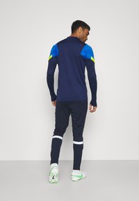 Marine trainingsjas met blauwe schouderaccenten, gecombineerd met marineblauwe broek met details in witte strepen. Witte sportschoenen met groene accenten.