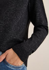 Czarny sweter o teksturowanej powierzchni z prążkowanymi mankietami, część codziennego stroju z jasnoniebieskimi dżinsami. Materiał ma delikatny połysk i subtelny wzór.