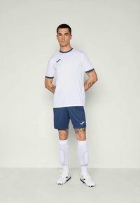 Biely dres s modrými akcentmi, doplnený modrými šortkami. Nosiť so bielymi ponožkami a futbalovými topánkami. Obsahuje logo na tričku a šortkách.