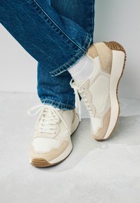 Zapatillas blancas con acentos de ante beige, suela de goma texturizada y diseño con cordones. Se llevan con vaqueros de mezclilla azul y calcetines blancos.