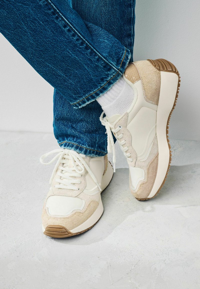 Zapatillas blancas con acentos de ante beige, suela de goma texturizada y diseño con cordones. Se llevan con vaqueros de mezclilla azul y calcetines blancos.