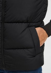 Schwarze gepolsterte Weste mit Reißverschluss vorne, seitlichen Taschen und gerippten Strickbündchen. Der Stoff ist glatt mit matter Oberfläche.