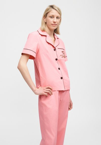 Blondynka w różowej, krótkim rękawem, zapinanej na guziki piżamie z czarnym obszyciem i pasujących różowych spodniach, stojąca z dłonią na biodrze.