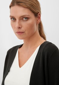 Zwarte cardigan over een witte V-hals top. De cardigan heeft een zachte, gestructureerde stof en een ontspannen ontwerp met een open voorkant en brede mouwen.