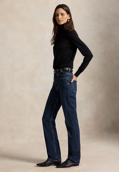 Femme debout, une main dans la poche, portant un col roulé noir, un jean bleu foncé, une ceinture noire et des chaussures noires sur un fond neutre.