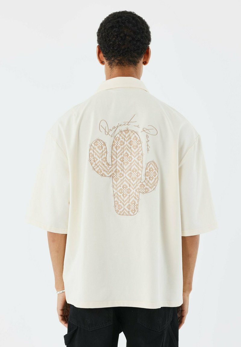 Camisa de manga corta color beige con un diseño texturizado de cactus y costuras decorativas en la parte posterior. Corte holgado con un cuello de solapa.