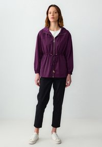 Vrouw die staat en een paarse zip-up jacket met taillekoord, zwarte broek, witte sneakers en een wit top draagt tegen een effen achtergrond.