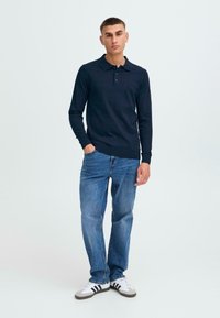 Marineblå langermet polo med tre knapper, kombinert med lyseblå jeans og hvite joggesko med svarte striper, stående foran en ensfarget bakgrunn.