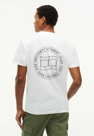 Man draagt een wit T-shirt met een rond Tommy Jeans-logo en tekst op de rug, gecombineerd met groene cargobroek.