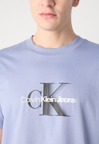 Hele lilla puuvillane t-särk ümmarguse kaelusega, millel on suur must-valge logo: "CK" ja "Calvin Klein Jeans" erksa tekstiga.