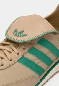 Μπεζ σουέντ αθλητικό παπούτσι με πράσινες λαστιχένιες λεπτομέρειες, που διαθέτει τρεις ρίγες και ραμμένη γλώσσα. Υλικά με υφή και αντιθετικά χρώματα.