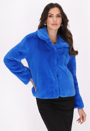 Frau mit langen dunklen Haaren, die eine hellblaue Kunstpelzjacke und einen schwarzen Rock trägt, steht mit einer Hand in der Hüfte.