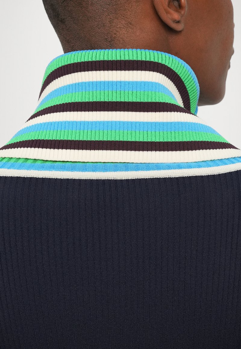 Geribbelde gebreide trui met een hoge kraag. Heeft afwisselende strepen in groen, blauw, wit en donkerbruin op een marineblauwe basis.