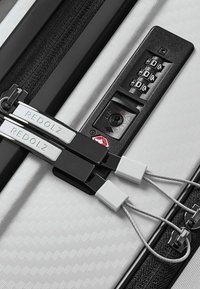 Primo piano di una valigia bianca texturizzata con lucchetto a combinazione nero e due cursori zip fissati da anelli in metallo.