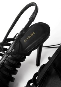 Sandali neri con tacco alto, dotati di sottili cinturini, finitura lucida e un motivo decorativo a incrocio. Design a stiletto con soletta imbottita.