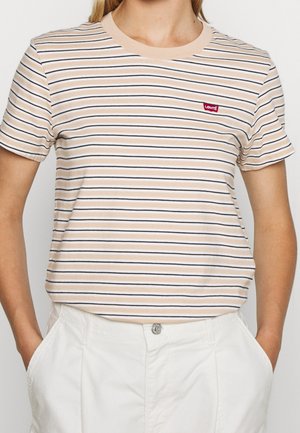 Personne portant un t-shirt Levi's beige et blanc à rayures, manches courtes, rentré dans un pantalon blanc, les mains dans les poches.