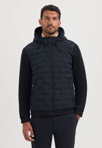 Gewatteerde zwarte jas met een capuchon, voorzien van een rits aan de voorkant, gebreide mouwen met structuur en twee zijzakken. Glad en gestructureerd materiaal gecombineerd.