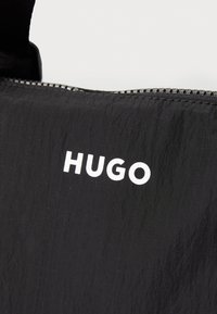 Czarna torba z tkaniny o teksturowanej powierzchni, z wyraźnym białym logiem "HUGO". Zamek błyskawiczny na górnej krawędzi, minimalistyczny design.
