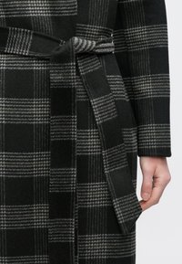 Casaco xadrez preto e cinza com tecido de tons duplos, apresentando um cinto na cintura e padrão texturizado, exibido com uma mão segurando o bolso lateral.