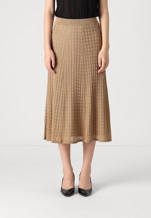 Vrouw draagt een beige geplooide midirok met een kabelgebreid patroon, zwarte top en zwarte puntige hakken, staand tegen een witte achtergrond.