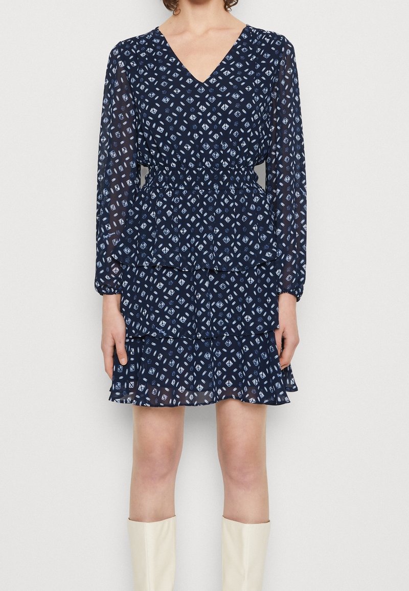 Námořnické modré šaty s dlouhými průhlednými rukávy, výstřihem do V, elastickým pasem a volánkovým lemem slušícím s bílým geometrickým potiskem po celém povrchu.