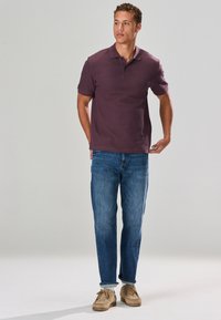 Polo de couleur violet foncé à manches courtes, associé à un jean en denim bleu. Des chaussures marron clair complètent le look, sur un fond neutre.
