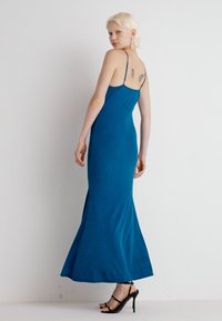 Vestido justos de comprimento total em teal com alças finas, textura ribada e uma abertura discreta nas costas, acompanhado de sandálias pretas de tiras.