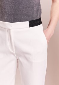 Pantalons blancs ajustés avec une finition texturée et des poches latérales. La ceinture élastique noire présente un détail scintillant subtil.
