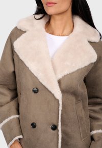 Brown double-breasted coat with a cream faux fur collar and trim. Features two black buttons and side pockets. Textured material, relaxed fit.