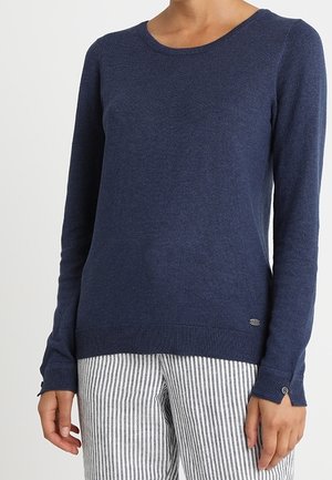 Femme portant un pull en maille bleu marine à manches longues associé à un pantalon à rayures verticales gris clair et blanc.