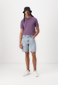 Polo shirt viola con un piccolo logo, pantaloni shorts di jeans blu chiaro con orlo sfrangiato e sneakers bianche. Cappello a secchiello nero con una trama liscia.