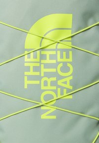 Light green fabric backpack with bright yellow logo and cord detailing. Features a textured surface and a distinctive graphic design.