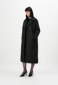 Woman with straight dark hair wears long quilted black coat, black semi-sheer tights, and black pointed mules with buckles, standing against white wall.