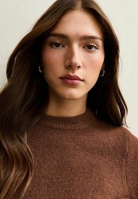 Bruine gebreide trui met een ronde hals, voorzien van geribbelde afwerking. Het haar is lang en golvend, waardoor het gezicht wordt omlijst tegen een neutrale achtergrond.
