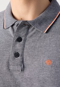 Gros plan d'une personne portant un polo gris texturé avec des boutons noirs et un bord de col rayé orange, avec un logo brodé orange.