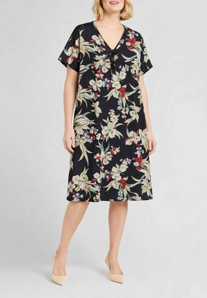 Donna che indossa un vestito nero a fiori fino al ginocchio, con maniche corte, scollo a V e tacchi alti beige, in piedi davanti a uno sfondo semplice.