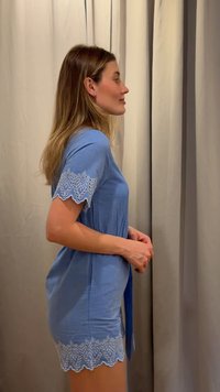 Oldalra fordulva álló nő, kék ruhában, fodros csipke ujjakkal és aljjal, bézs függöny háttér előtt.