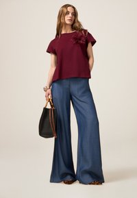 Top bordeaux a maniche corte con accento floreale, abbinato a pantaloni in denim blu a gamba larga e una borsa nera a righe con manici marroni.