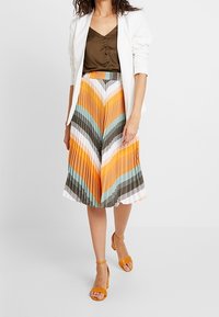 Falda midi plisada con un patrón de chevron en tonos naranja, turquesa y marrón, combinada con una blazer blanca y un top marrón. Tacones amarillos con tiras.