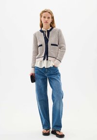 Knit cardigan in beige with navy accents, round neck, front pockets, paired with light blue straight-leg jeans and black flip-flops.