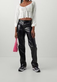 Žena v bílém vypasovaném topu s proříznutým vzorem, lesklých černých texturovaných kalhotách, černo-bílých teniskách a s malou růžovou kabelkou.