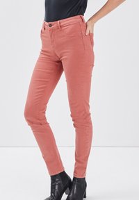 Vaqueros ajustados de color rosa con una textura suave, cinco bolsillos y un cierre de botón en la cintura. Combinados con botines negros.