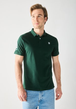 Camisa polo de algodón verde con cuello, dos botones y un logo en la manga izquierda; combinada con jeans de color azul claro sobre un fondo liso.