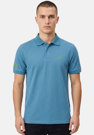Uomo che indossa una polo blu a maniche corte con loghi geometrici verdi sul petto e sulla manica sinistra, su uno sfondo semplice.