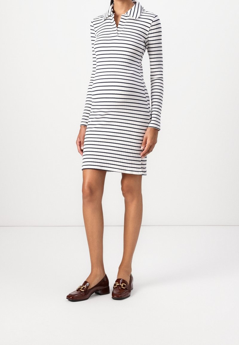 Femme portant une robe blanche côtelée avec des rayures horizontales noires et des manches longues, assortie à des mocassins en cuir marron avec des boucles dorées.