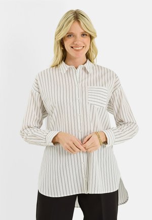 Jeune femme souriante portant une chemise blanche à boutons avec de fines rayures noires verticales et une poche avant, associée à un pantalon noir.