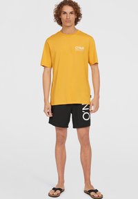 Gelbes Baumwoll-T-Shirt mit kurzen Ärmeln und weißem "O'Neill"-Logo. Kombiniert mit schwarzen Badeshorts mit weißer Schrift und schlichten Sandalen.