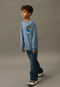 Svijetloplava dukserica s grafikonom Sonica; dugi rukavi i rebrasti manžeti. Nositi s tamnoplavim trapericama i sivim tenisicama.